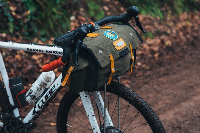 Muddy Gravel Amateur with a canvas Carradice bag strapped to the front on a DIY support, on a leafy dirt track.