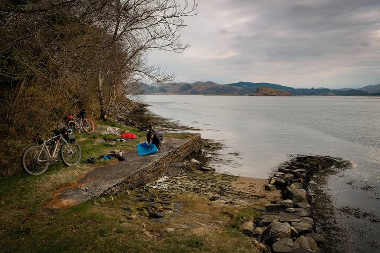 Bikepacking bivvy spot on a stone slipway beside the sea at Crinan, with two loaded bikes and a rider packing kit.