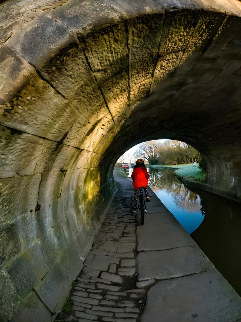 Child in a red coat rides a bike along a cobbled canal towpath through a stone bridge near Macclesfield.