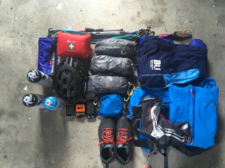 Bikepacking kit laid out on a concrete floor: helmet, first aid kit, dry bags, bottles, GPS units, shoes and clothing.