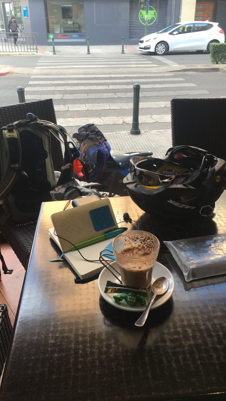 Cafe table with a hot drink, open notebook, helmet and rucksack, a loaded bike parked behind on a town street.