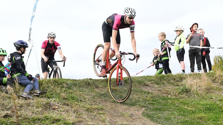 Rider on the red Cube Cross Race Pro drops off a grassy bank during a cyclocross race, spectators watching.