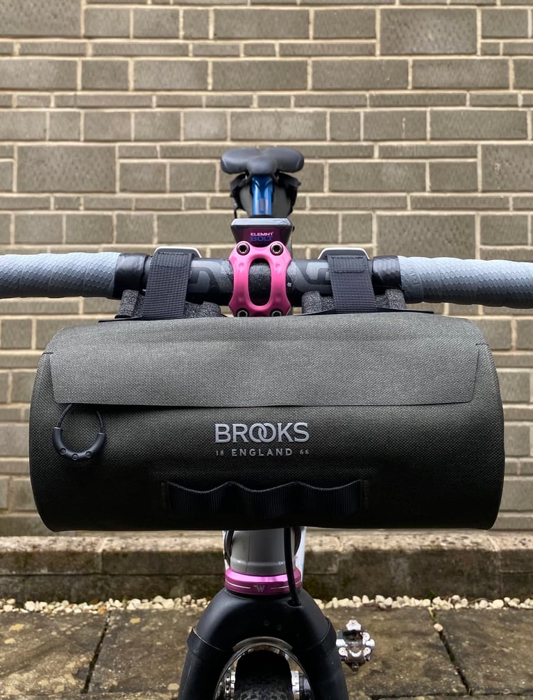 Brooks Scape Handlebar Pouch mounted on drop bars, seen head-on against a brick wall, with a pink stem above it.