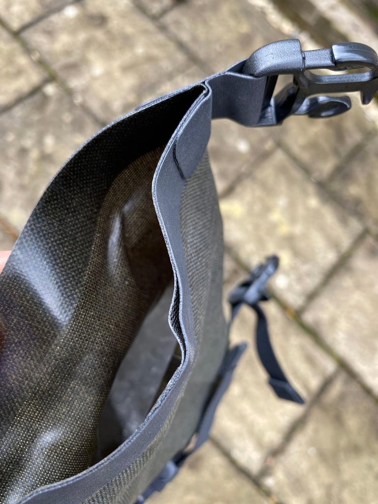 Open roll-top end of the Brooks Scape Handlebar Roll dry bag held over paving slabs, with its buckle clip at the corner.