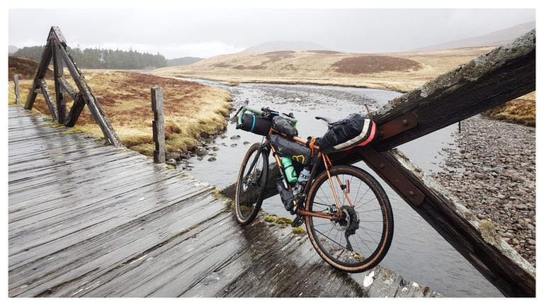 Loaded orange gravel bike leant on a wet timber bridge over the River Pattack, Central Highlander route, Scotland.