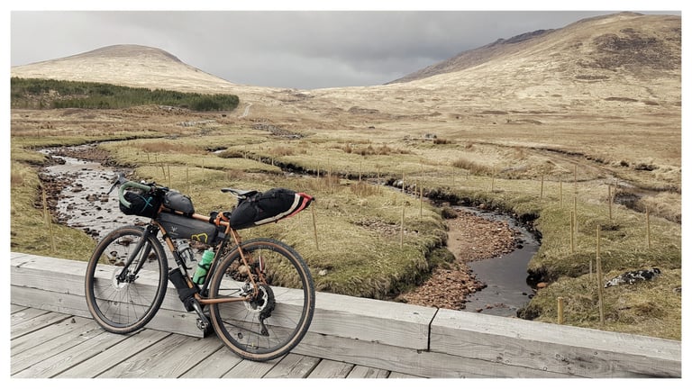 Bikepacking bike propped on a timber bridge over a small burn with rounded hills behind, Central Highlander route.