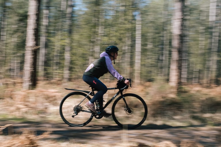 Rider in Rapha All Day Leggings on a gravel bike in motion, with blurred pine trees behind.