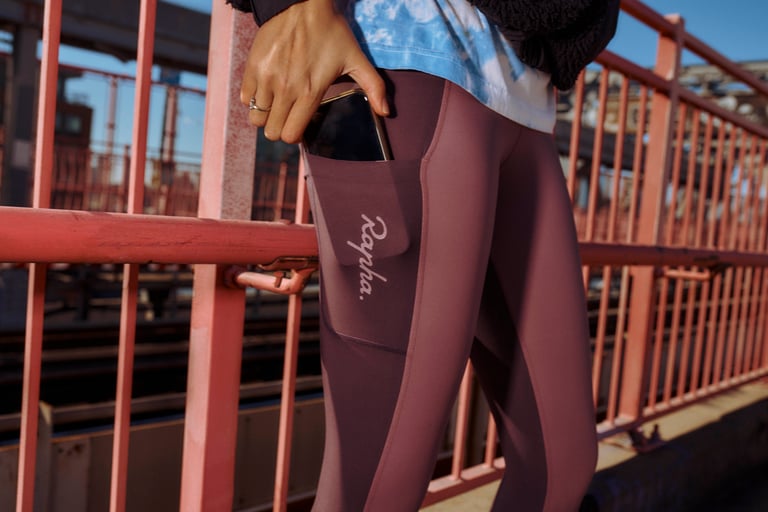 Hand slips a phone into the thigh pocket of two-tone purple Rapha All Day Leggings on a pink-railed bridge.