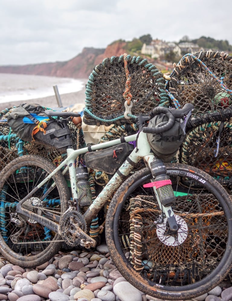 Muddy Canyon Grizl in Matcha Splash paint leaning on lobster pots on a pebble beach in Devon.
