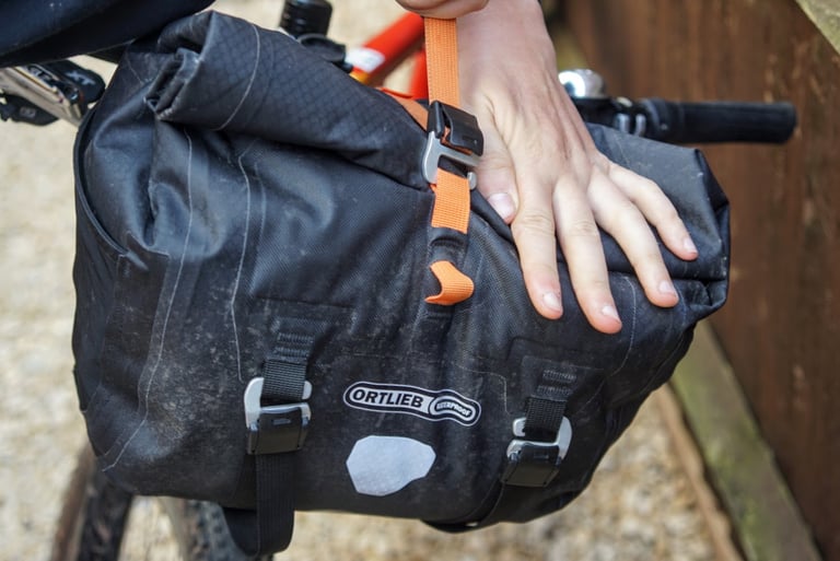 Hand presses the rolled top of the Ortlieb Handlebar Pack QR, secured by an orange strap and CamLock buckle.