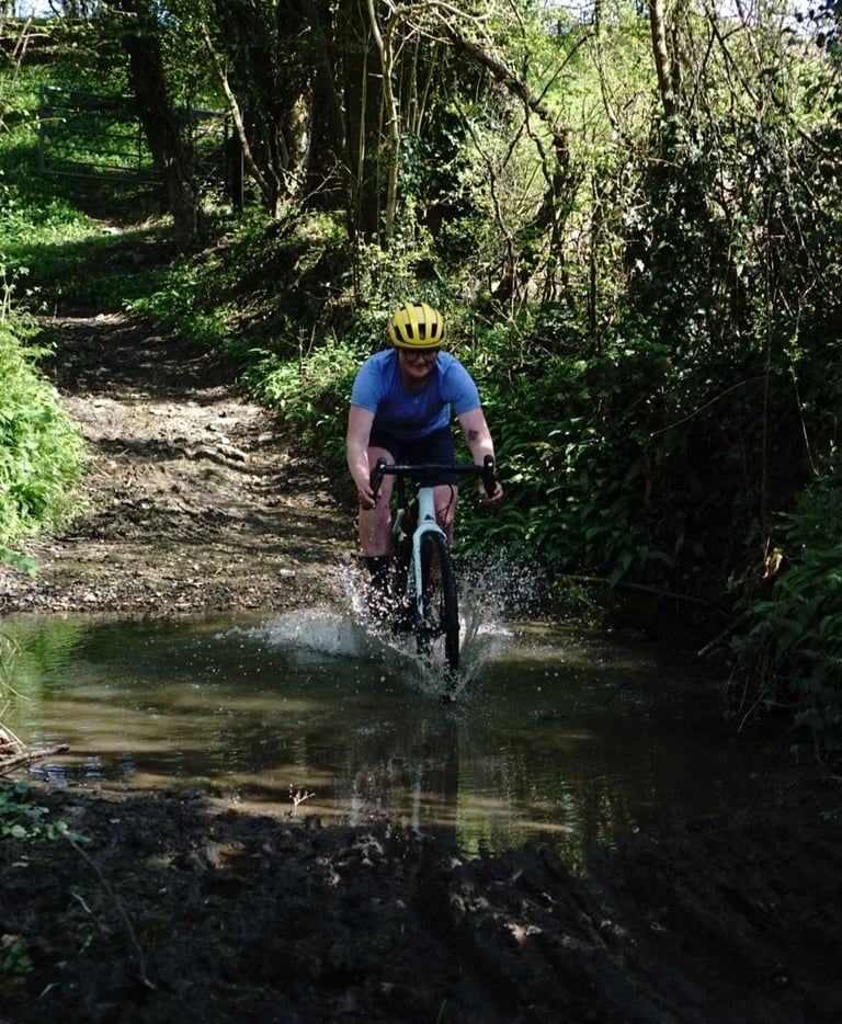 Rider in a yellow helmet splashes through a muddy ford on the Canyon Grizl during the review.