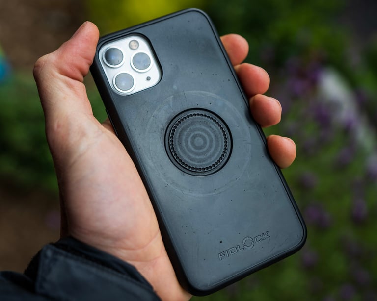 Hand holding a black Fidlock VACUUM phone case on an iPhone 11 Pro, showing the round mount plate on the back.