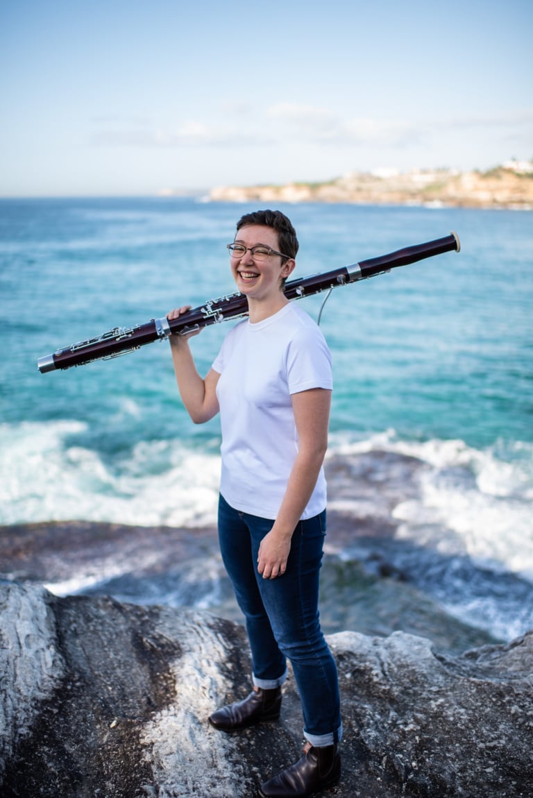 Alison Wormell, performer of the SOLOS I bassoon recital films, stands on sea rocks with her bassoon over one shoulder.