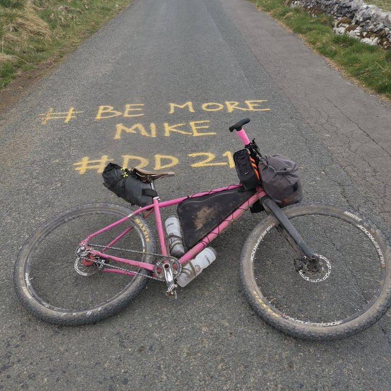Pink bikepacking bike lying on a Dales lane beside chalked road text reading #BE MORE MIKE #DD21.