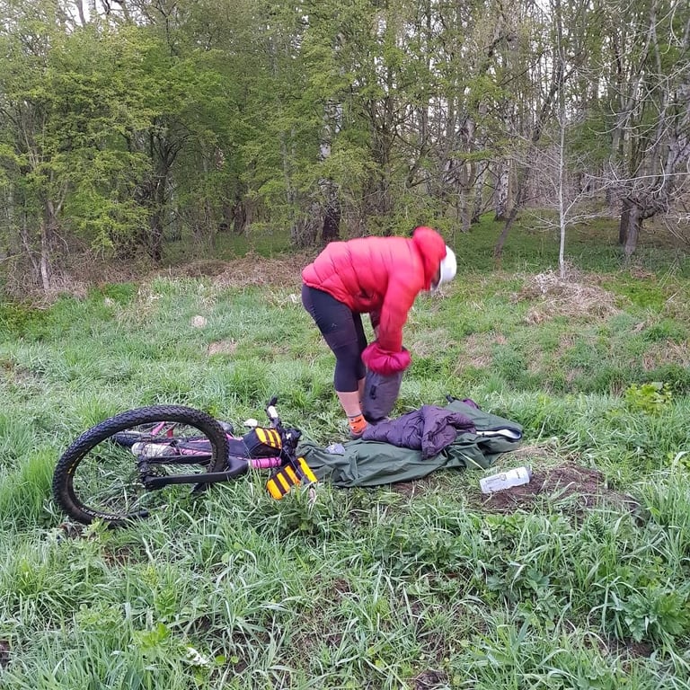 Rider in a red down jacket packs up a damp bivvy camp at the woodland edge, a pink bikepacking bike lying in the grass.