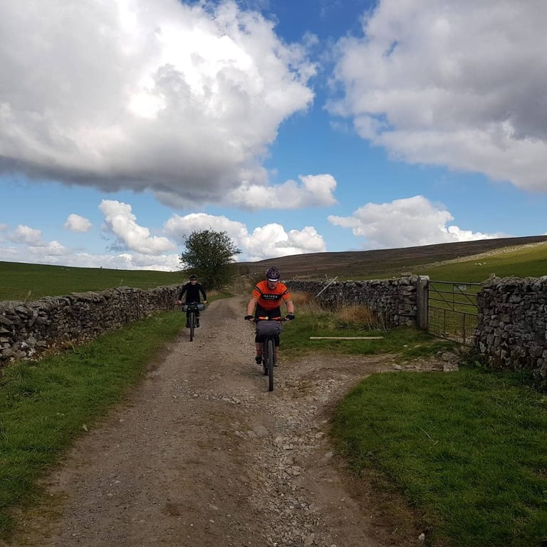 Two Dales Divide riders descend a rocky farm track between dry stone walls under a big sky, one in an orange jersey.