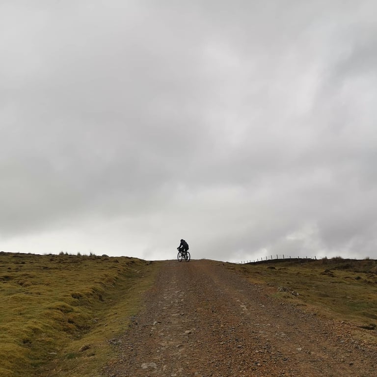 Stu crests a stony moorland climb on the Dales Divide, silhouetted against heavy grey cloud.