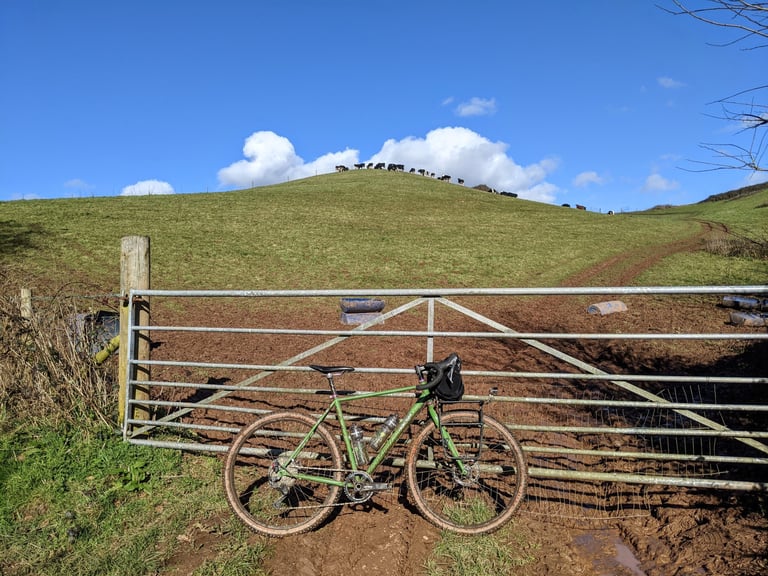Green gravel bike with Ortlieb Handlebar Pack QR on drop bars, leaning on a muddy farm gate below a hill.