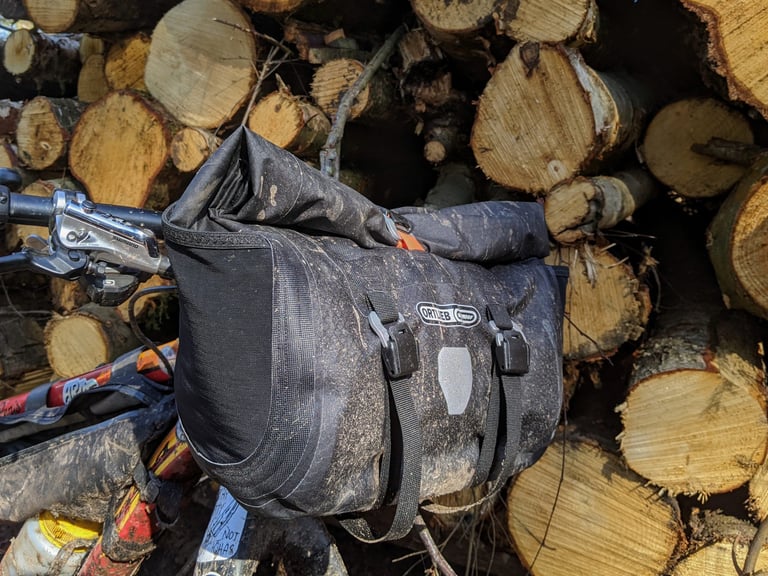 Muddy Ortlieb Handlebar Pack QR on a bike in front of a log pile, with side pocket and buckled straps.