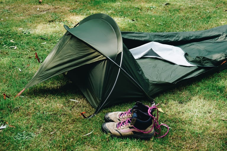 Alpkit Elan hooped bivvy pitched on grass with the hood open and a pair of walking boots in front of it.