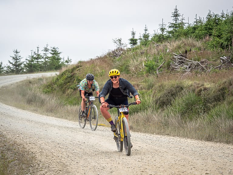 Smiling Gritfest rider on a yellow bike with race number 225 on a gravel forest track, another rider close behind.