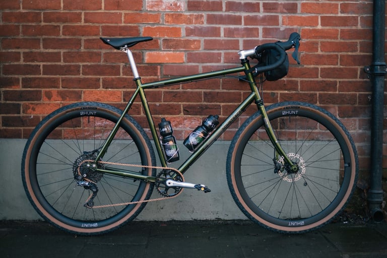 Bombtrack Arise built by The Woods Cyclery, in green with Hunt wheels and tan-wall Teravail tyres, against a brick wall.