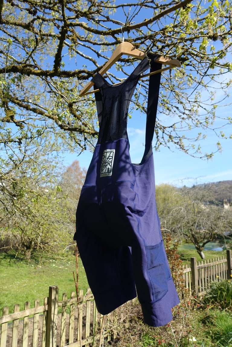 Victory Chimp Out There Cargo Bib Shorts in navy hanging from a wooden hanger in a tree, with a perforated mesh back.