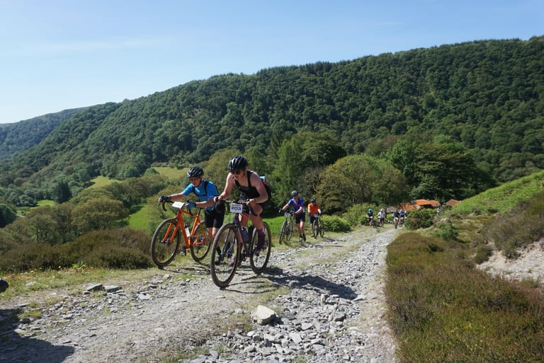 Gritfest riders push their bikes up the steep, rocky K2 climb in Mid Wales, with others strung out behind.