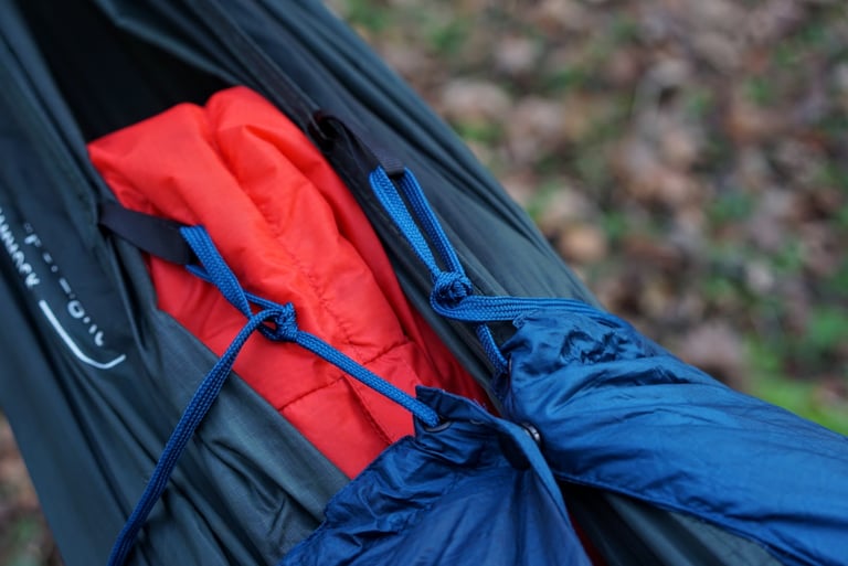 Blue cords from the Cloud Cover quilt tied to the tabs of a DD hammock, with a red sleeping bag inside.