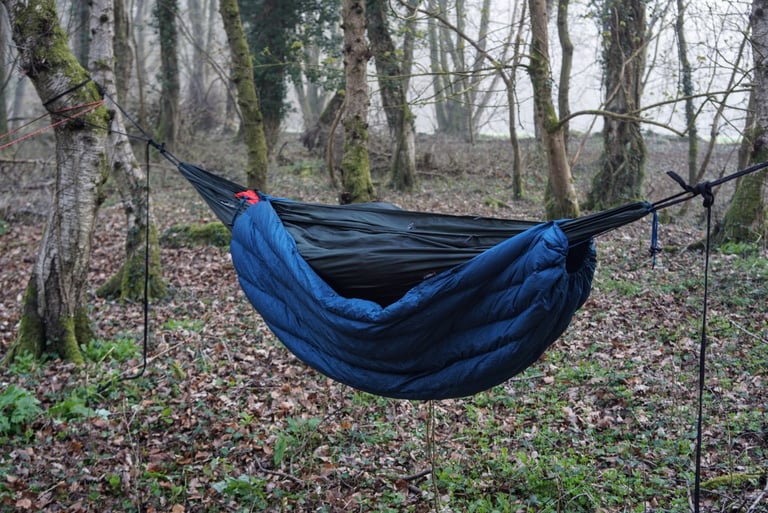 Blue Alpkit Cloud Cover quilt rigged beneath a dark green hammock as an underblanket in misty woodland.