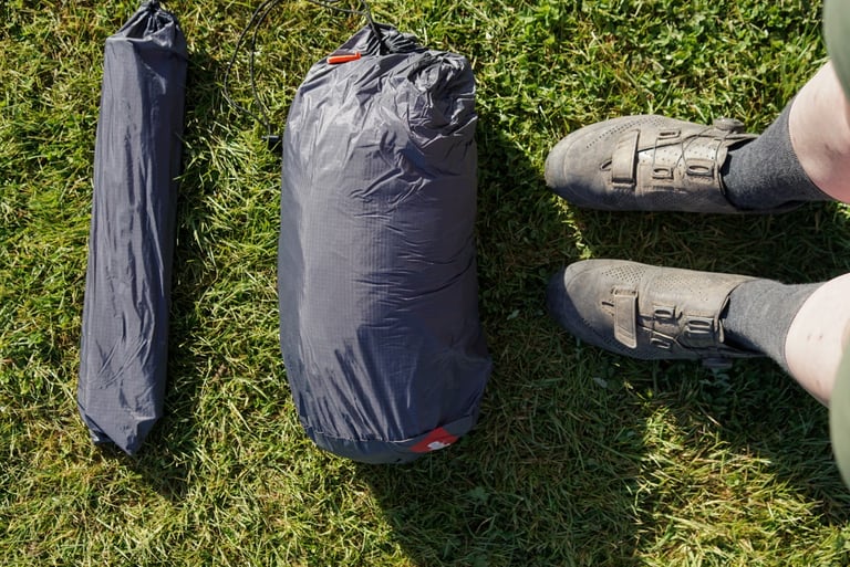 Packed Alpkit Jaran 2 tent bag and slim pole bag on grass next to a rider's cycling shoes for scale.