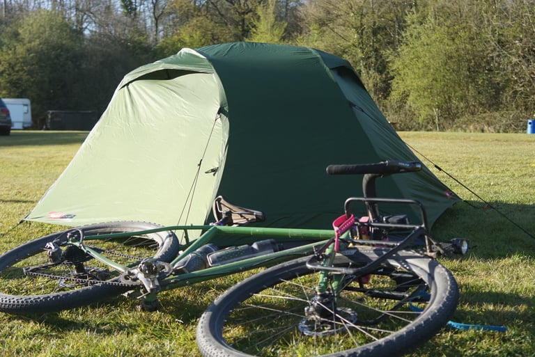 Green Alpkit Jaran 2 tent pitched on a campsite with a bike with a front rack lying on the grass in front of it.