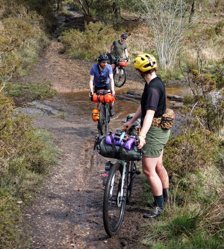Rider in Machines For Freedom Key Shorts wheels a loaded bike through a muddy ford as two bikepackers ride across.