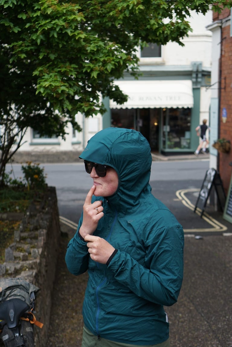 Rider in the Velocio Ultralight Hooded Jacket and sunglasses pauses on a village street opposite a cafe.