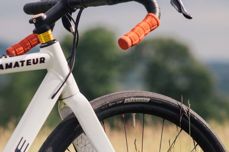 Front end of a white Cube cross bike with orange grips and a Teravail Rampart 700c x 38 tyre in front of a green field.