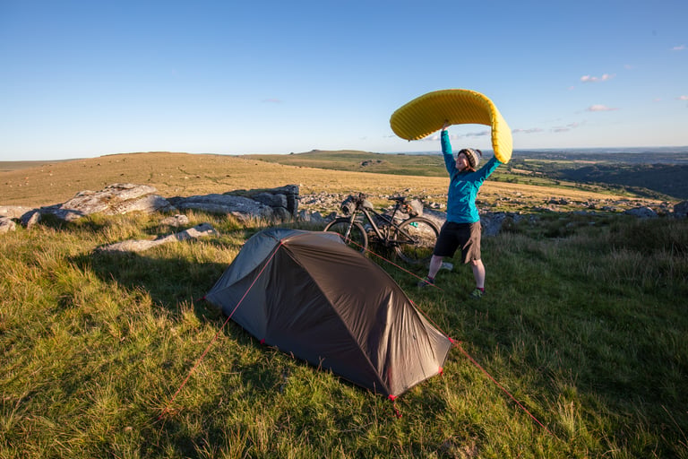 Bikepacker holds a yellow sleeping mat aloft beside a tent and bike while wild camping on Dartmoor.