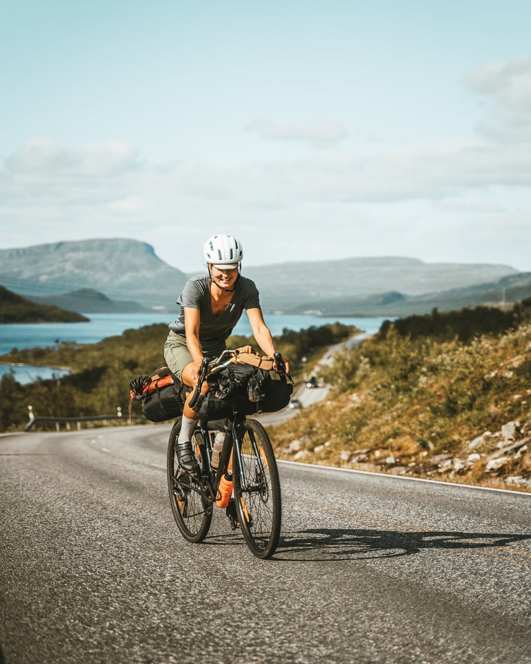 Henna rides a loaded gravel bike along a lakeside road in Finland for her Cycling Through A Pandemic story.