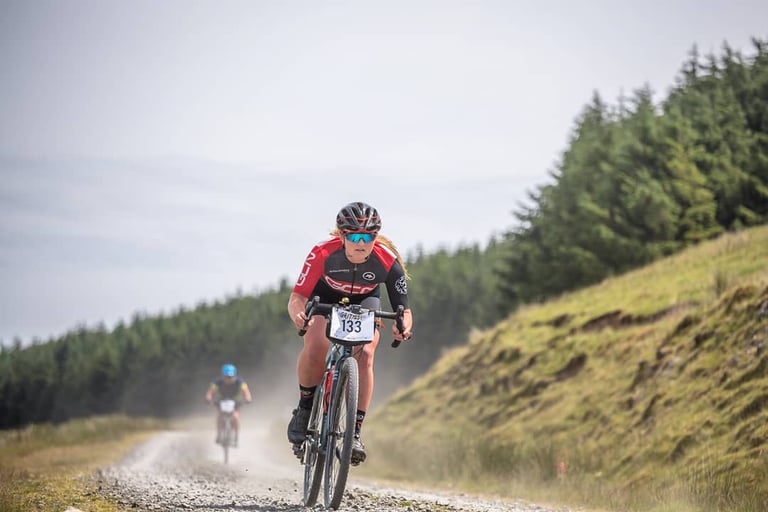 Gritfest rider with number 133 on the bars rides a dusty gravel track beside a conifer plantation, another rider behind.
