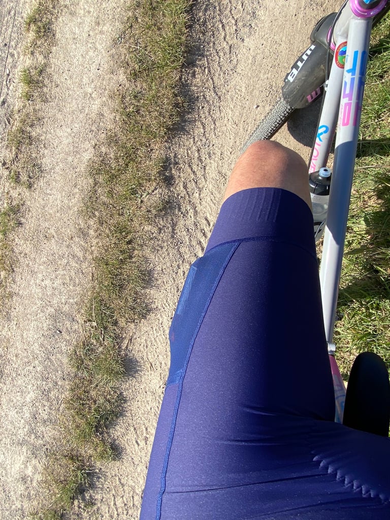 Rider's thigh in navy Victory Chimp Out There bib shorts beside a gravel bike on a sandy track, showing the leg gripper.