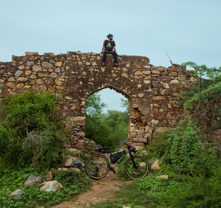 Rider sits on top of a ruined stone archway in the jungle in India, a bikepacking mountain bike parked below.