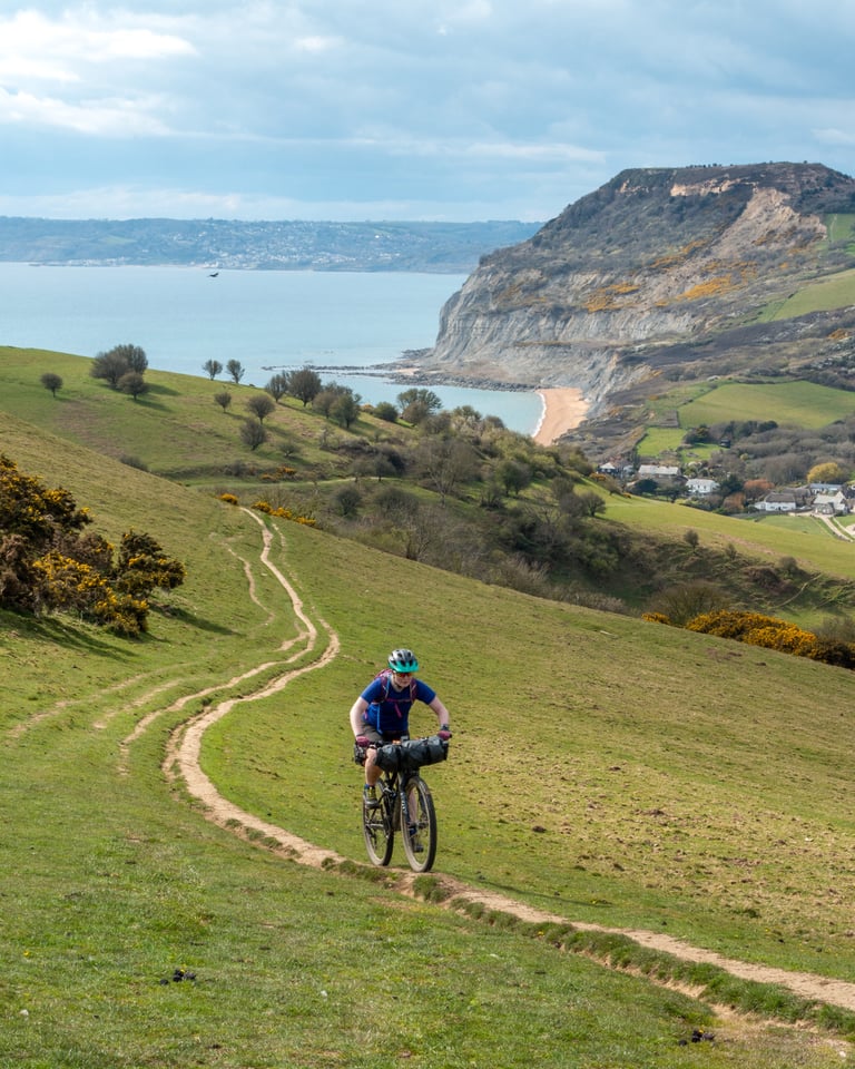 Bikepacker climbs a grassy track above the Jurassic Coast, with cliffs and sea behind.