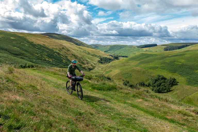 Emma Kingston rides a mountain bike with a handlebar bag along a grassy hillside above a green valley.