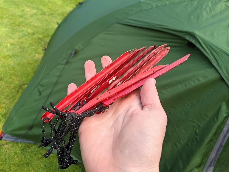 Hand holding the ten red Alpkit pegs from the Jaran 2 tent, with black reflective cords and the green flysheet behind.