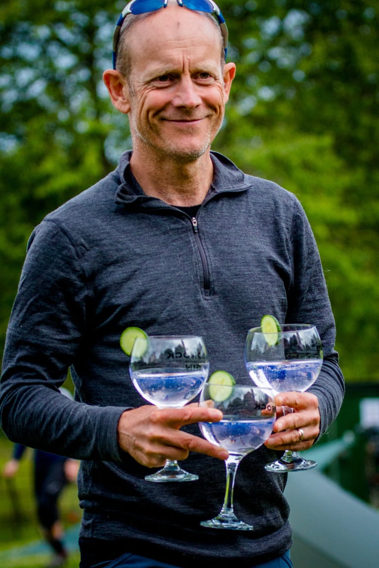 Smiling man in a grey half-zip carries three gin and tonics garnished with cucumber at The Distance.