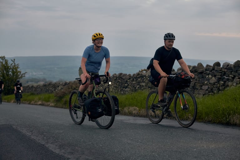 Two riders on a Yorkshire lane, one with Restrap panniers on a Tubus Tara front rack, beside a dry-stone wall.