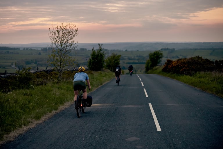 Three riders on a quiet road at dusk, the nearest carrying Restrap panniers, with fields stretching behind.