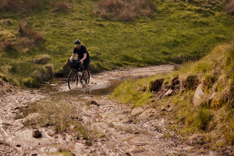 Jon Hicken splashes a Fairlight Secan 2 with a handlebar bag through a muddy stream on a moorland track.