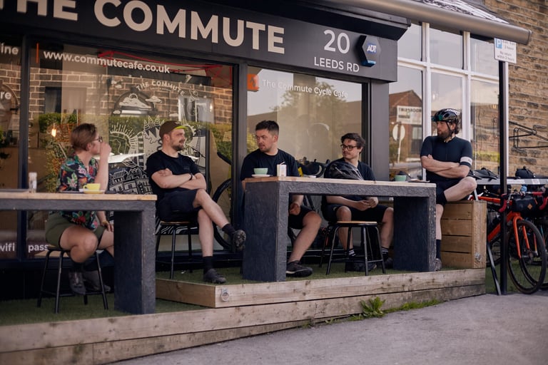 Five riders sit at outdoor tables outside The Commute Cycle Cafe on Leeds Road, Ilkley, bikes parked beside them.