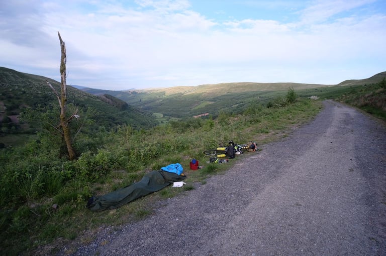Simple bivvy bag laid out beside a gravel track above a forested valley, with a loaded bikepacking bike nearby.