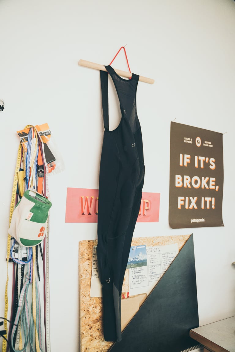Black cycling bibs hang in a repair workshop beside a poster reading 'If it's broke, fix it', for the Velocio repair scheme.